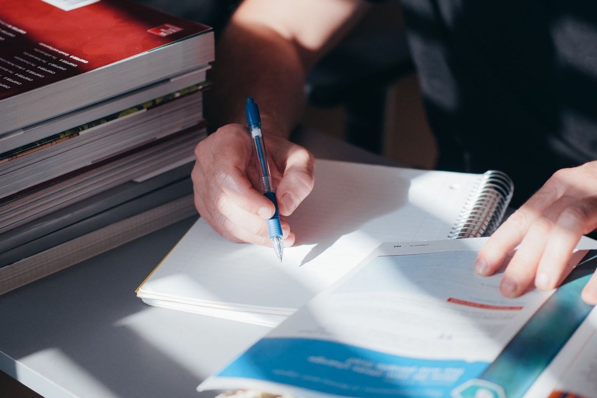 Una persona toma apuntes en un cuaderno junto a una pila de libros